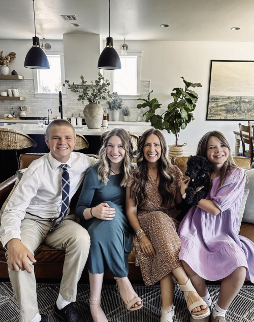 A family of four sits on a couch in a modern living room. Two women, a man, and a girl holding a black dog are smiling at the camera. The room has a white and beige color scheme with plants and pendant lights in the background.